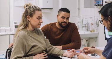 A pregnant patient receives a glucose test while speaking with a healthcare professional alongside her partner.