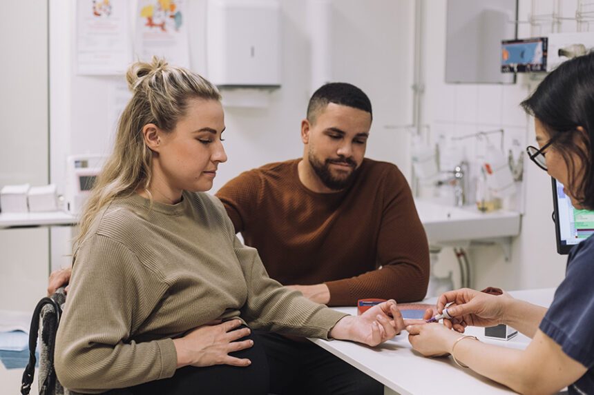 A pregnant patient receives a glucose test while speaking with a healthcare professional alongside her partner.