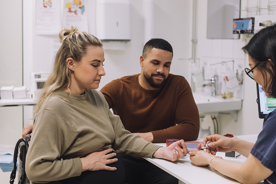 A pregnant patient receives a glucose test while speaking with a healthcare professional alongside her partner.