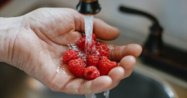 A person rinses fresh raspberries under running water in a kitchen sink.