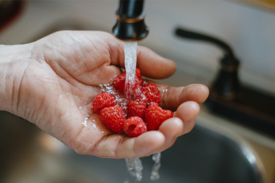 A person rinses fresh raspberries under running water in a kitchen sink.