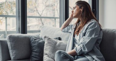A woman sits on a couch looking thoughtfully out the window.