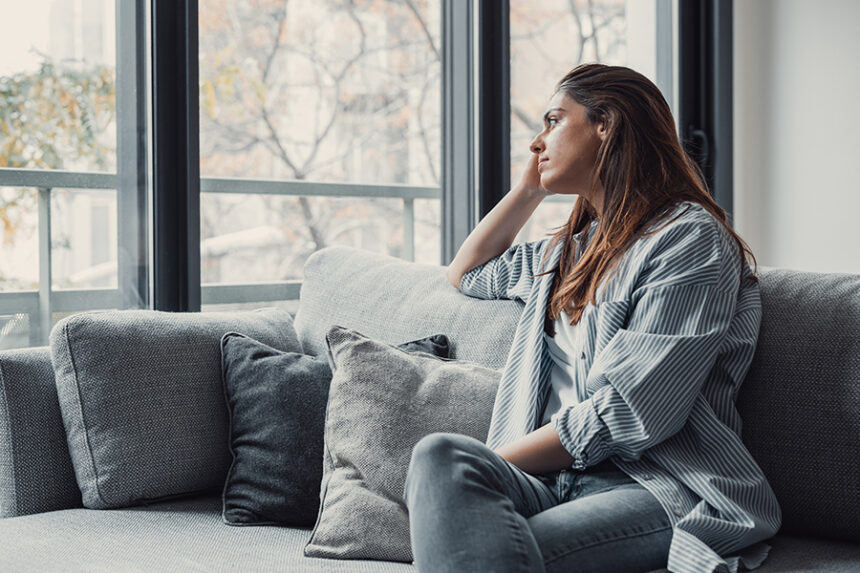 A woman sits on a couch looking thoughtfully out the window.