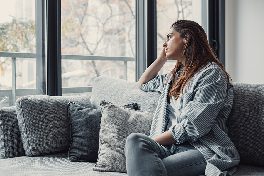 A woman sits on a couch looking thoughtfully out the window.