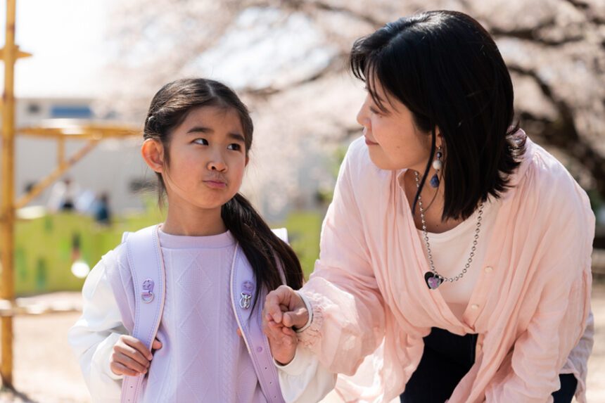 A mother talks with her young daughter outside as the child wears a backpack for school.