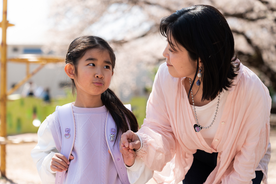 A mother talks with her young daughter outside as the child wears a backpack for school.