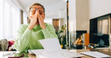 A woman sits at a table looking stressed while reviewing bills and paperwork.
