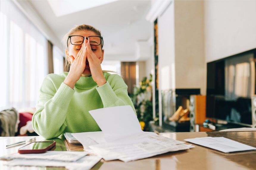 A woman sits at a table looking stressed while reviewing bills and paperwork.