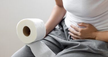 Woman holding a roll of toilet paper and clutching her abdomen while sitting on a toilet.