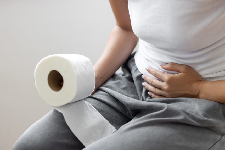 Woman holding a roll of toilet paper and clutching her abdomen while sitting on a toilet.
