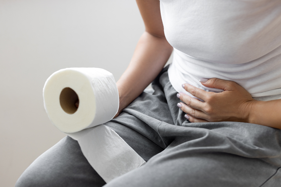 Woman holding a roll of toilet paper and clutching her abdomen while sitting on a toilet.