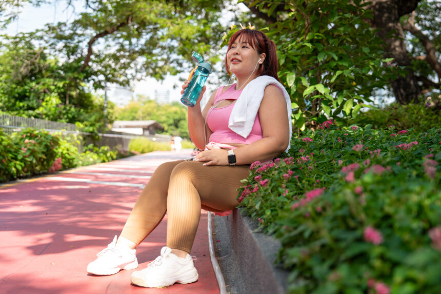 woman stops to take a drink of water on a bench while walking in a park